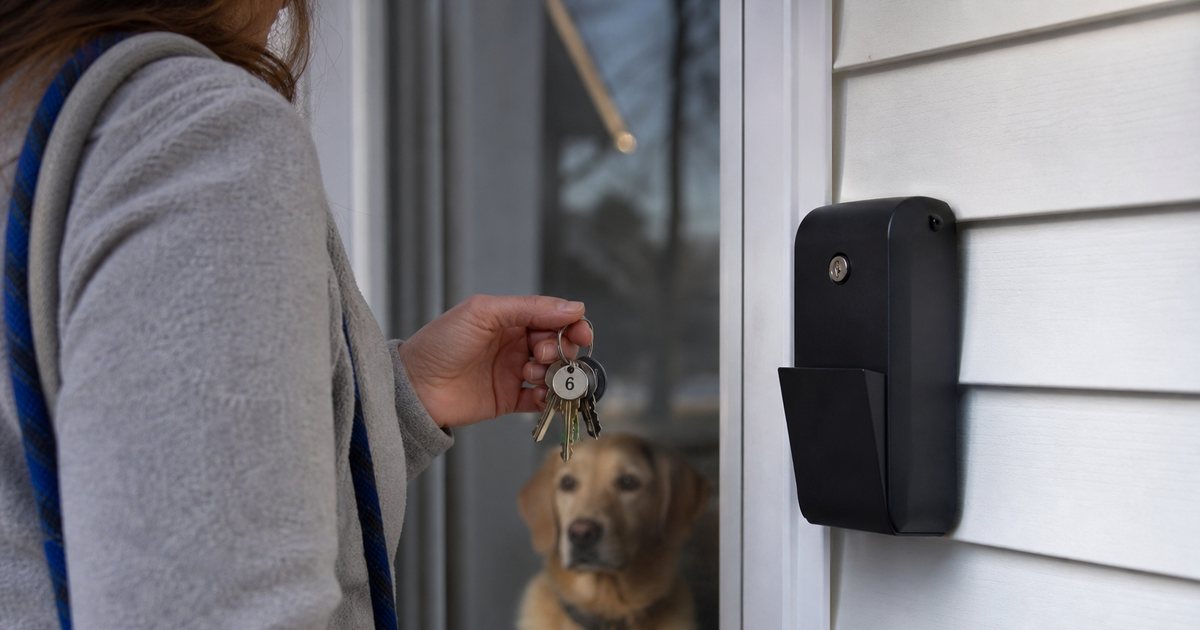 A dog walker's hand holding a small ring of keys with plain numbered tags beside a wall mounted lockbox on a front porch, a leash over the shoulder and a dog waiting behind the storm door, soft morning light